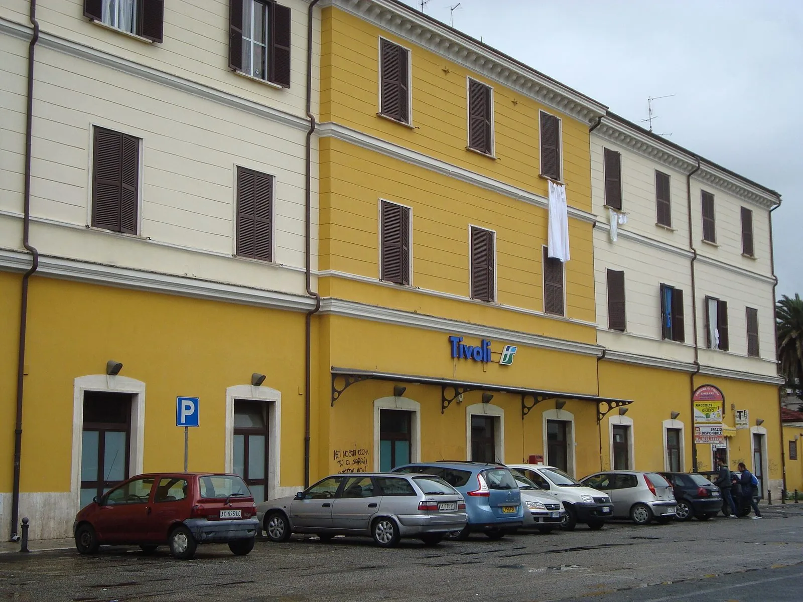 Facciata gialla e bianca della stazione ferroviaria di Tivoli con l'insegna e alcune auto parcheggiate davanti