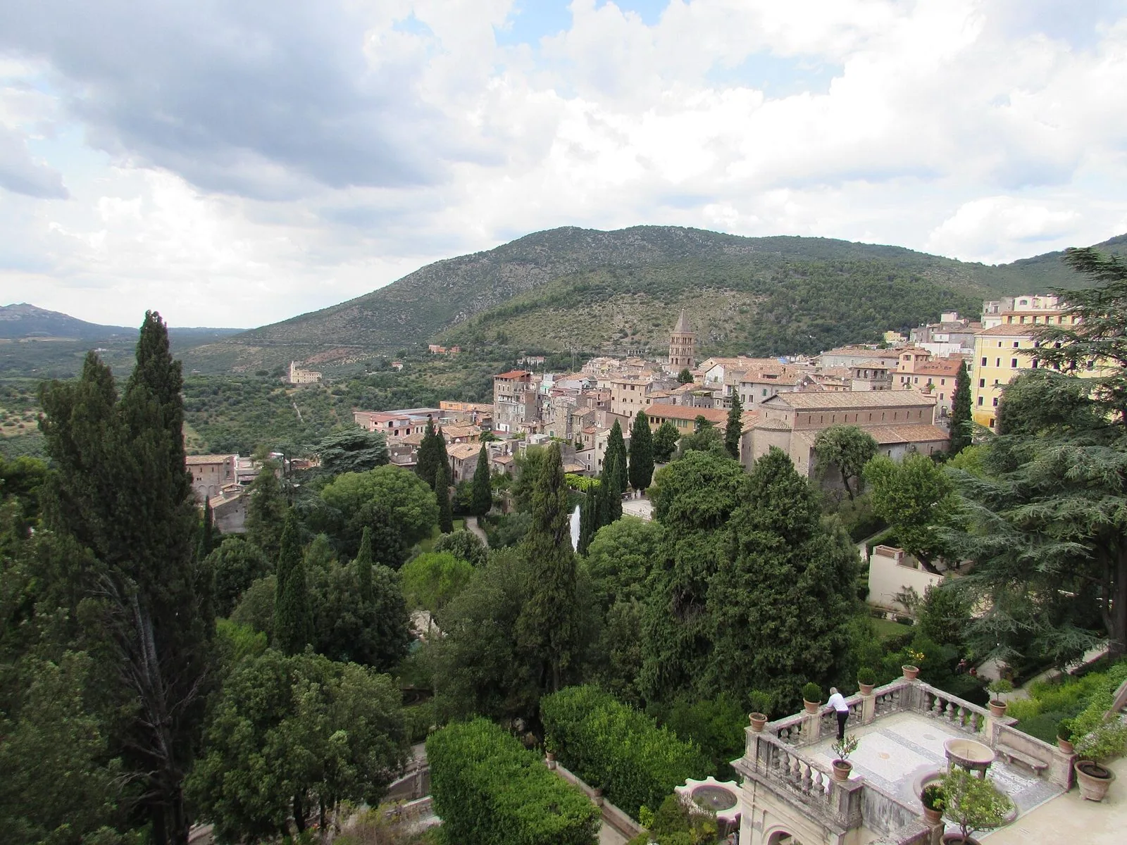 Veduta di Tivoli dai giardini di Villa d'Este: cipressi e alberi in primo piano, il centro storico con un campanile romanico e i Monti Tiburtini sotto un cielo nuvoloso